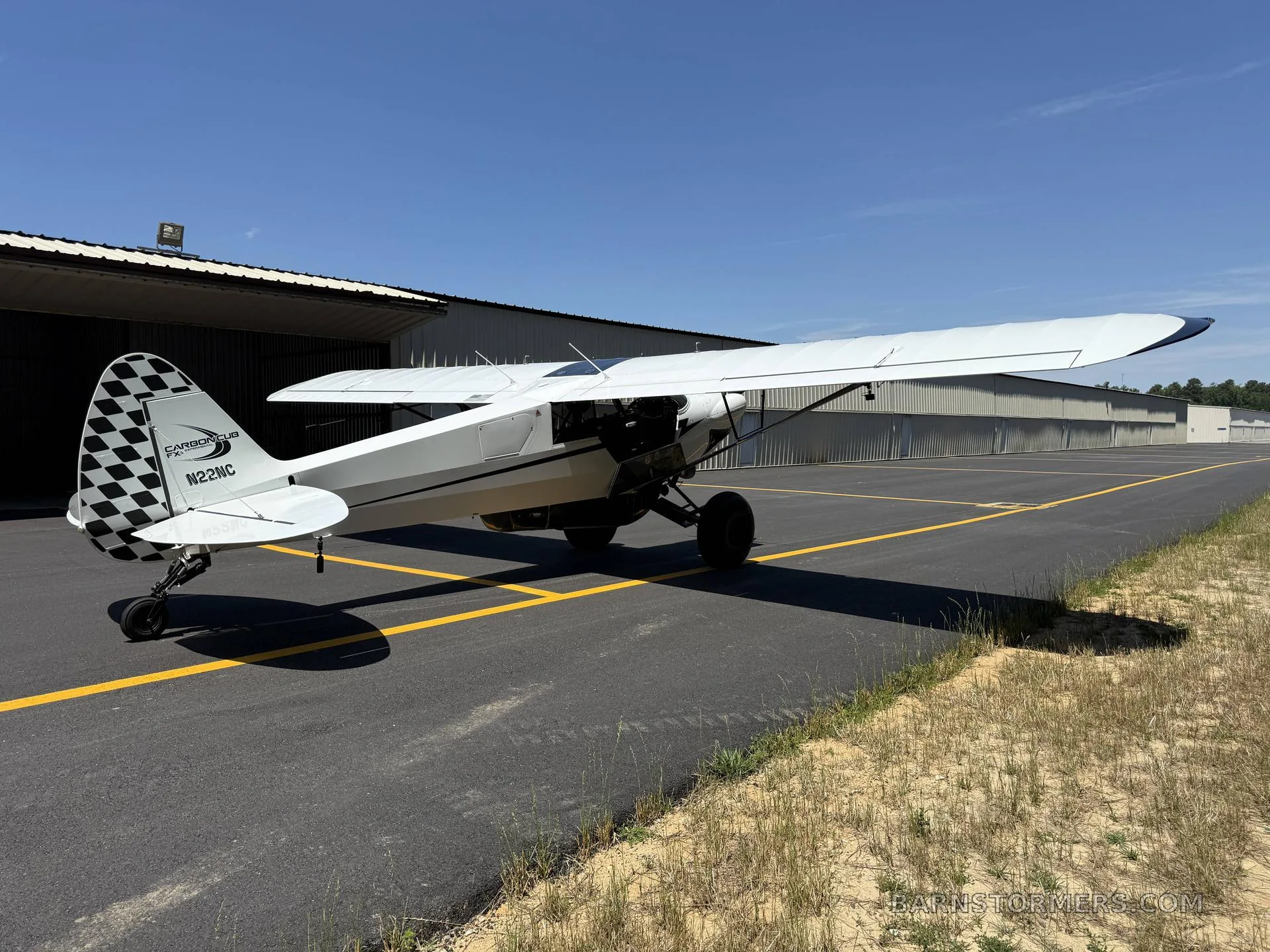 A rear-quarter view of a white Carbon Cub FX-3 aircraft with a checkered tail is parked on an asphalt ramp next to a hangar.