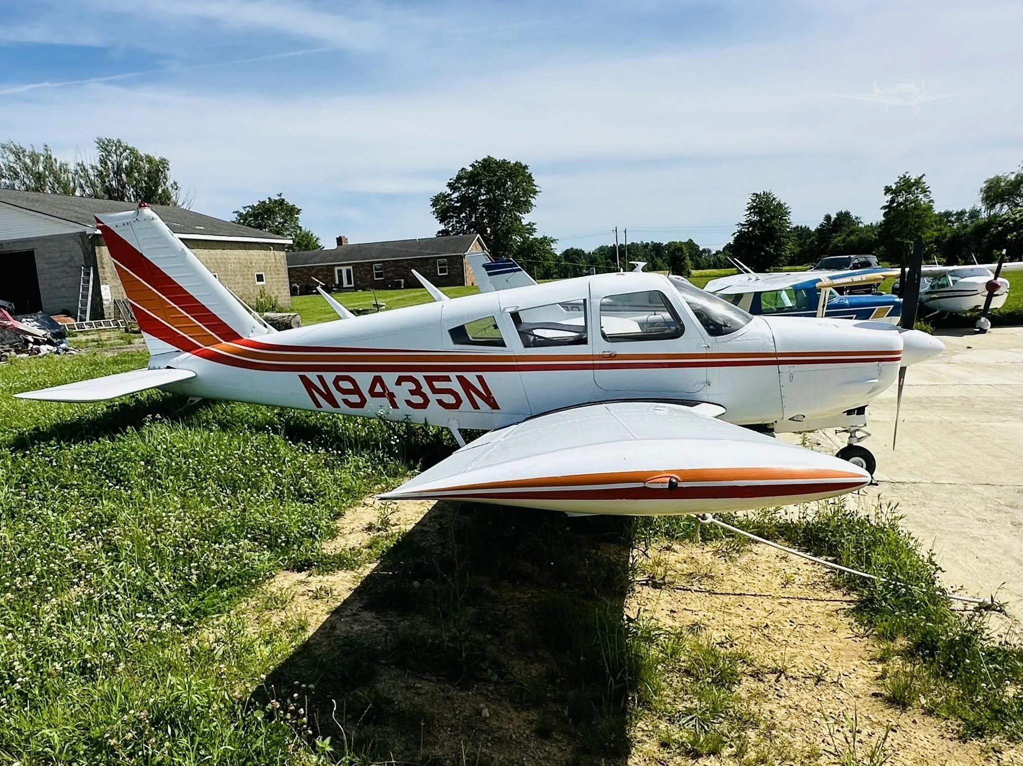 A white single-engine propeller aircraft with red and orange stripes, registration N9435N, parked on grass next to a concrete pad under a blue sky.