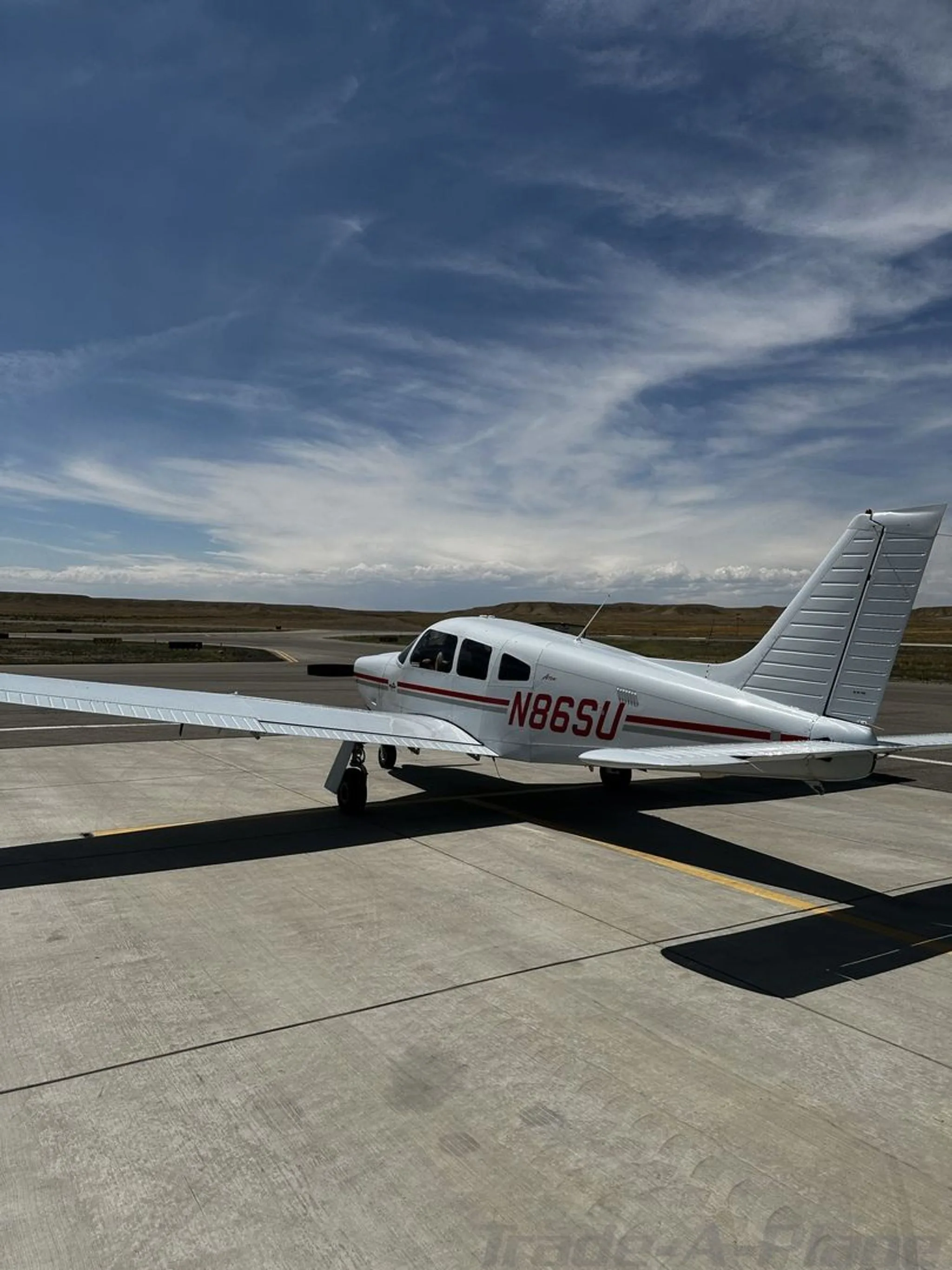 A white Piper aircraft with red stripes and tail number N86SU is parked on a concrete tarmac under a blue sky.