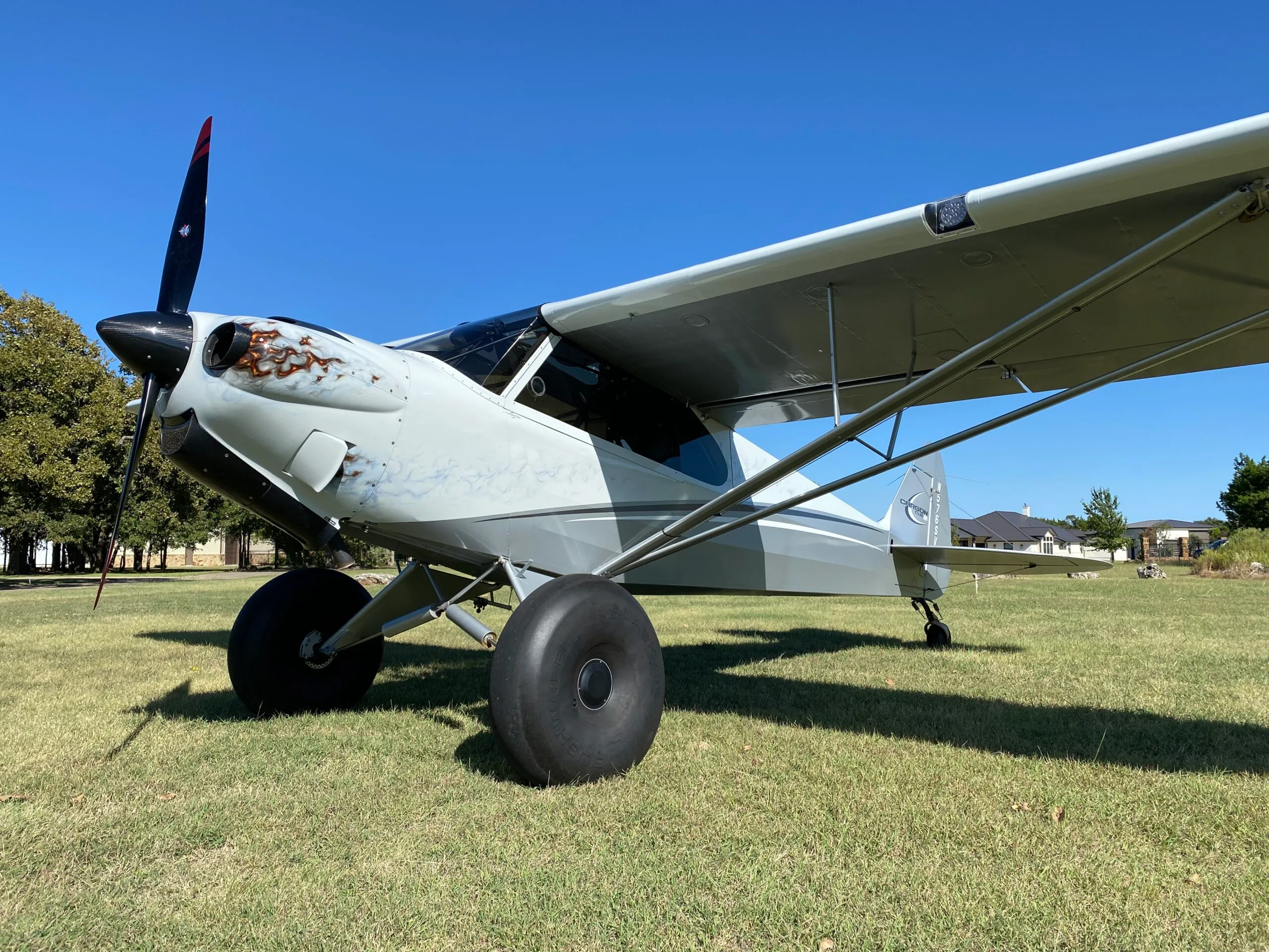 A light grey Carbon Cub aircraft with a unique flame-like paint scheme on the cowling and large bush tires, parked on a grassy field under a clear blue sky.