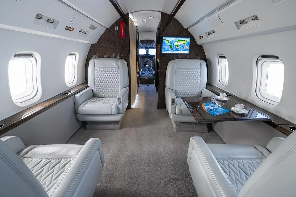 The luxurious interior cabin of a business jet features light gray leather seats and a table with coffee cups, looking towards the cockpit.