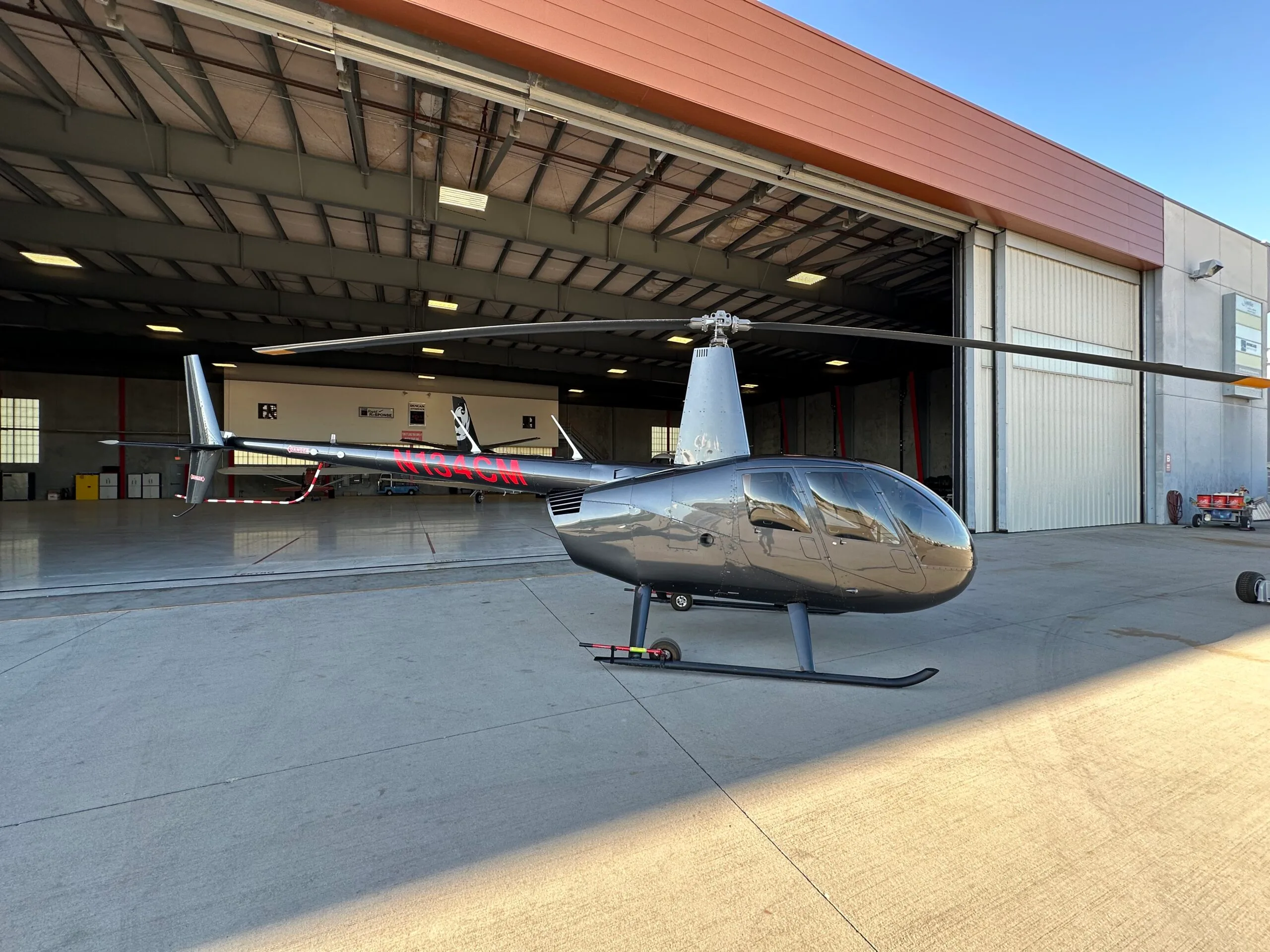 A dark grey Robinson R44 helicopter with red registration N184CM is parked on a concrete apron in front of an open hangar.