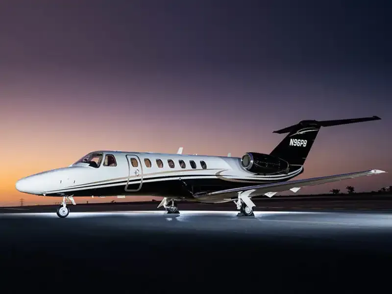 A white and black Cessna Citation business jet with gold stripes on a tarmac at dusk, illuminated by ground lights.