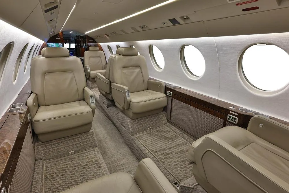 A perspective view of the business jet's cabin, featuring beige leather seating and large windows.