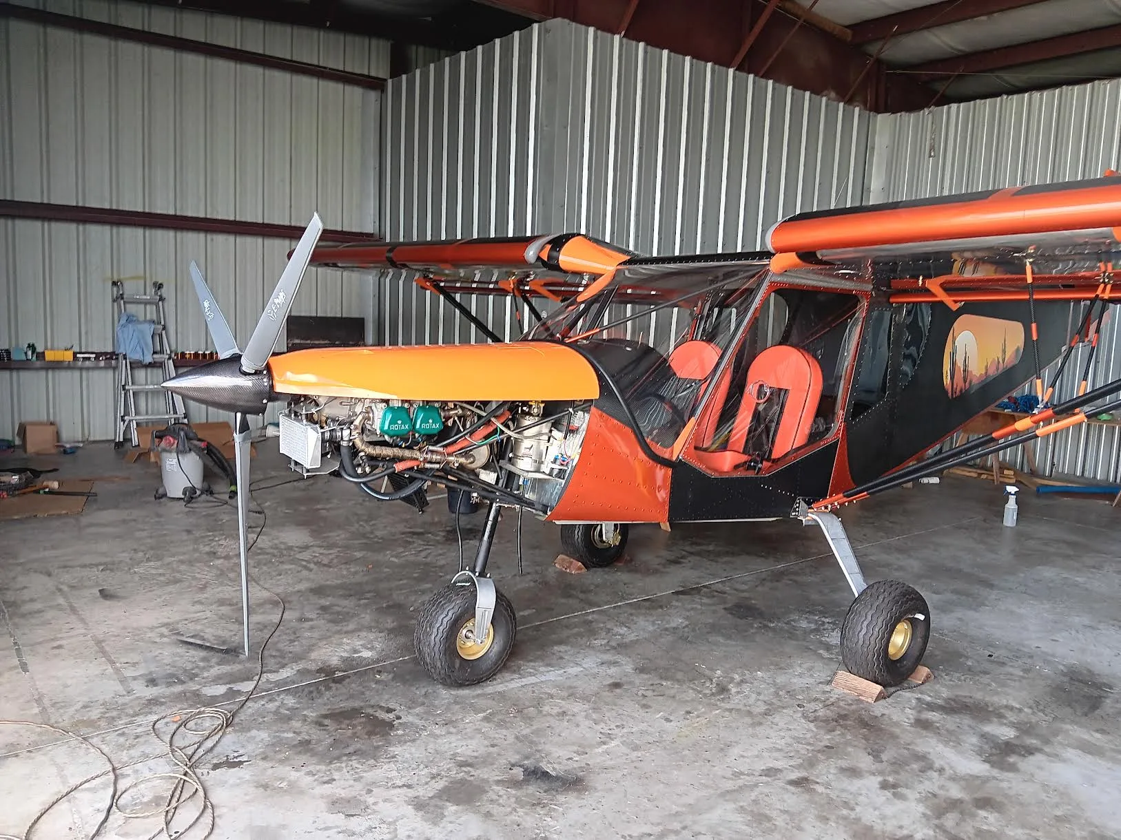 An orange and black ultralight aircraft with an exposed engine and desert sunset mural, parked inside a hangar.