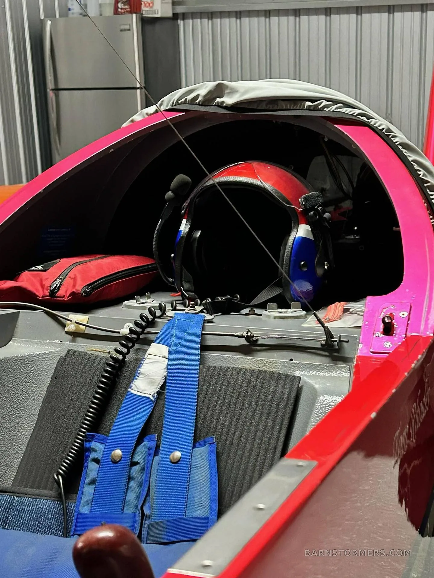 The interior of an aerobatic aircraft, showing a red and blue helmet, a red bag, and blue harnesses in the storage area behind the seat.
