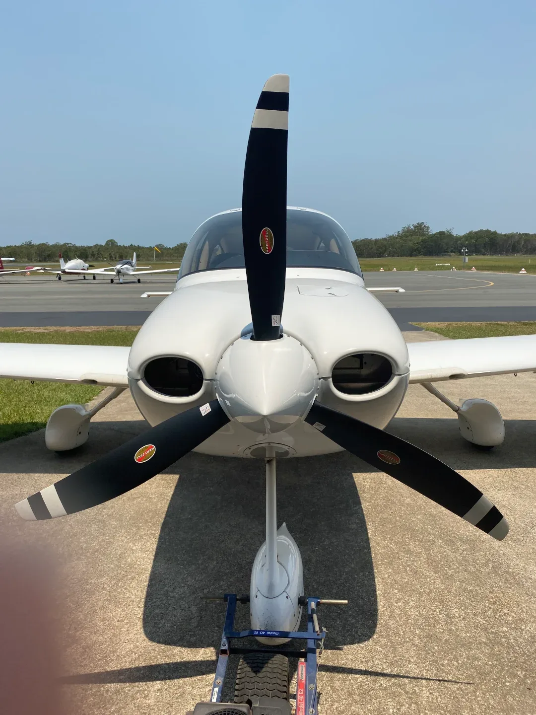 A white single-engine propeller aircraft is parked on an airfield, viewed from the front with other planes in the background.
