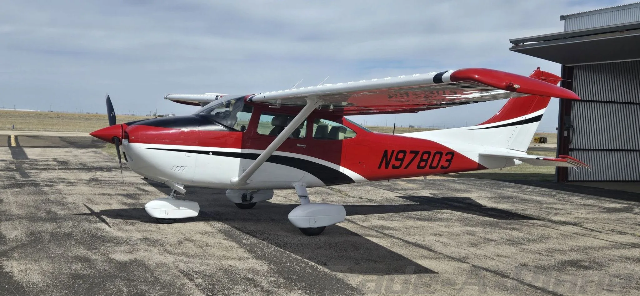 A red, white, and black Cessna aircraft (N97803) is parked on an airfield next to a hangar.