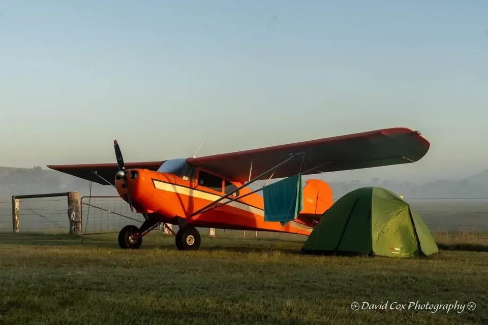 An orange and white high-wing aircraft parked next to a green tent in a grassy field during a misty morning.