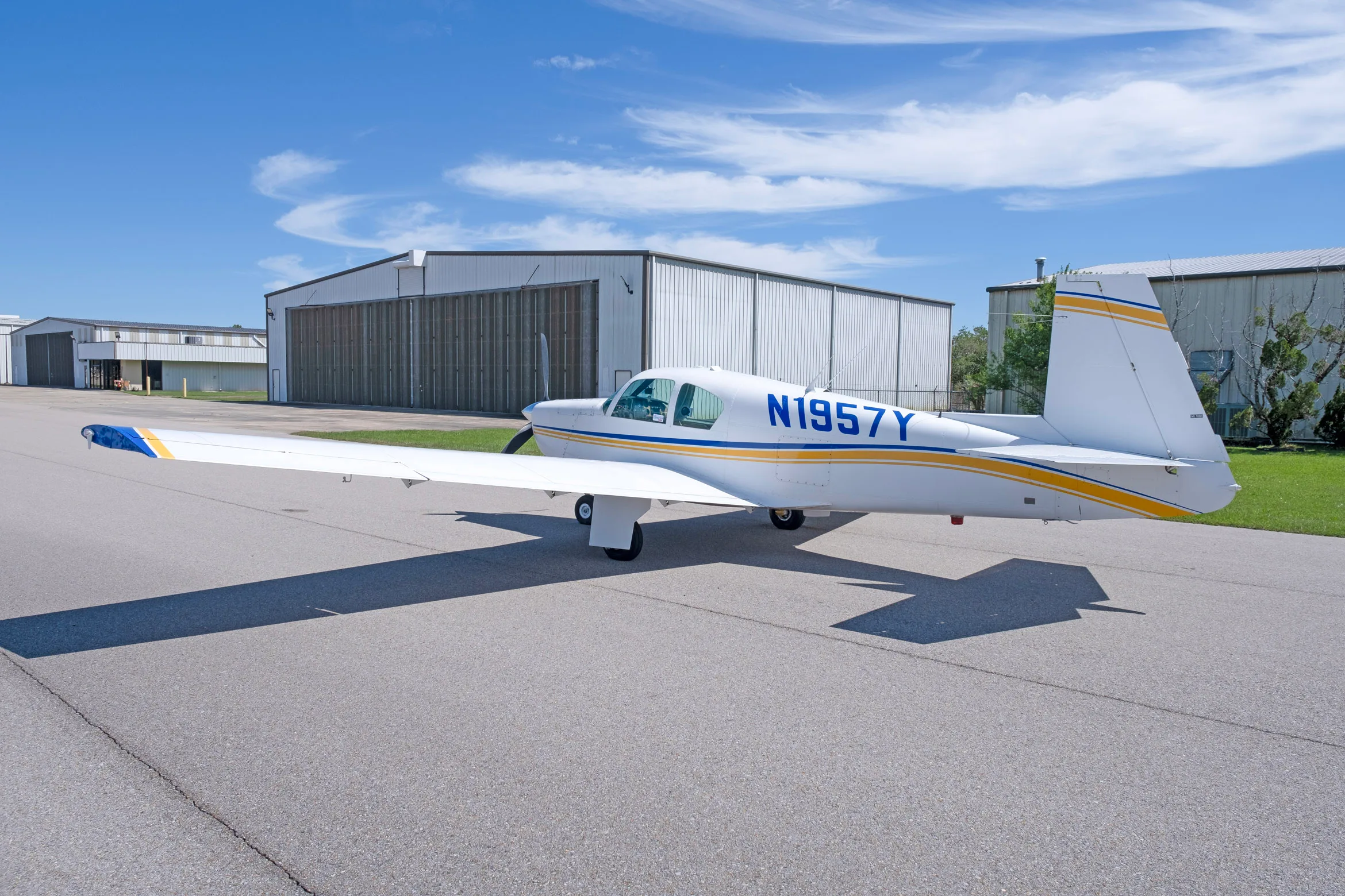 A rear-side view of a white single-engine propeller aircraft with blue and yellow stripes, registration N1957Y, parked on an asphalt apron.