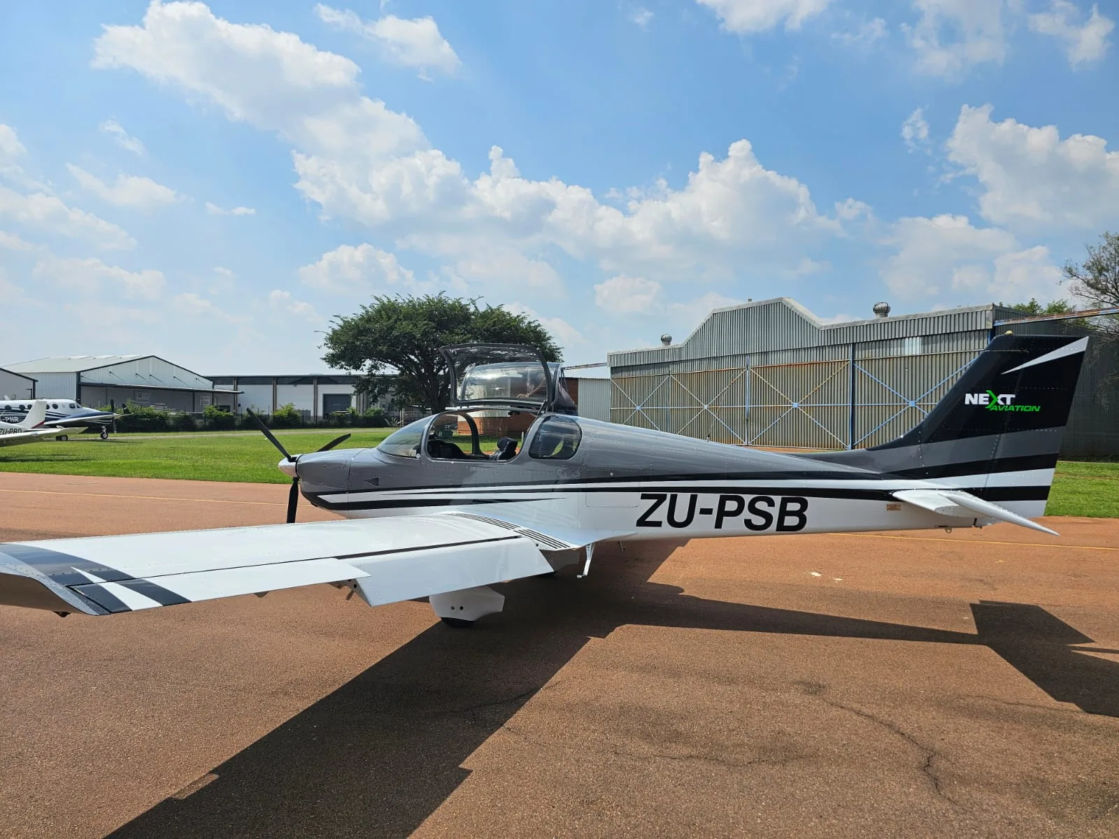 A grey and white single-engine propeller aircraft, ZU-PSB, with its canopy open, parked on a tarmac with hangars in the background.