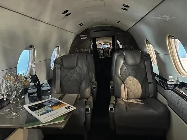 The luxurious cabin interior of a business jet with two grey leather seats, a table set with drinks, and large windows.