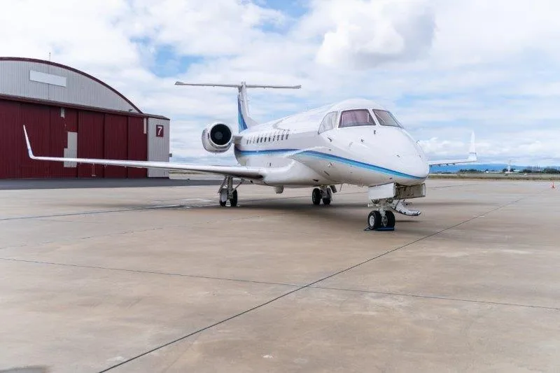 A white business jet with blue stripes is parked on an airfield tarmac in front of a red hangar under a cloudy sky.