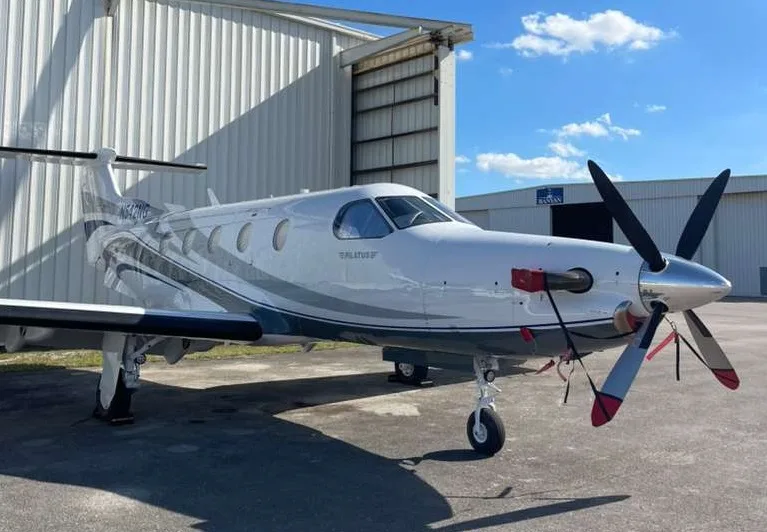 A white Pilatus PC-12 NG aircraft with blue and grey stripes, registration N542NG, parked in front of a hangar.