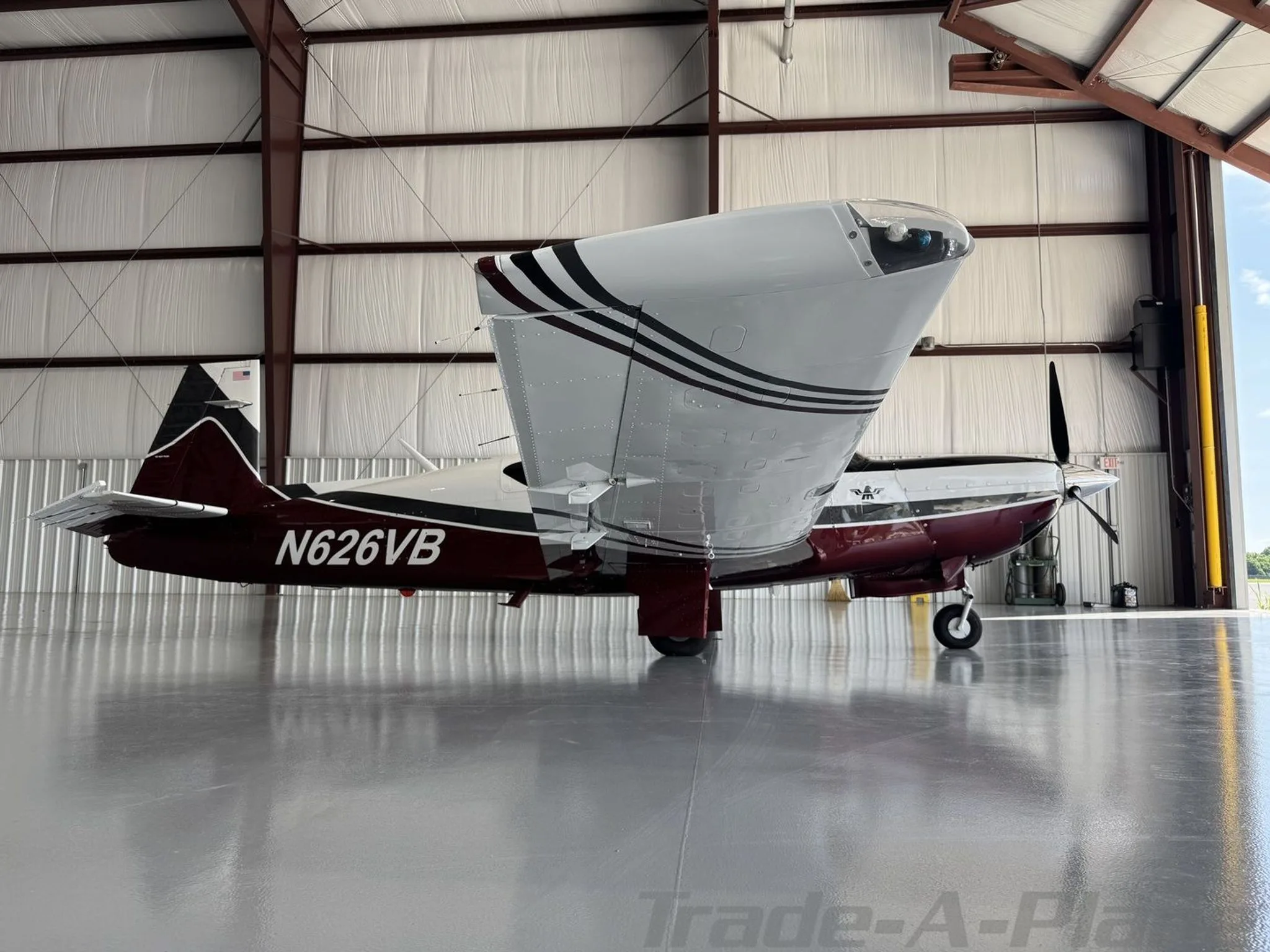 A Mooney aircraft with registration N626VB, featuring a maroon, white, and black paint scheme, is parked in a hangar with the door open, viewed from the side showing the underside of the wing.