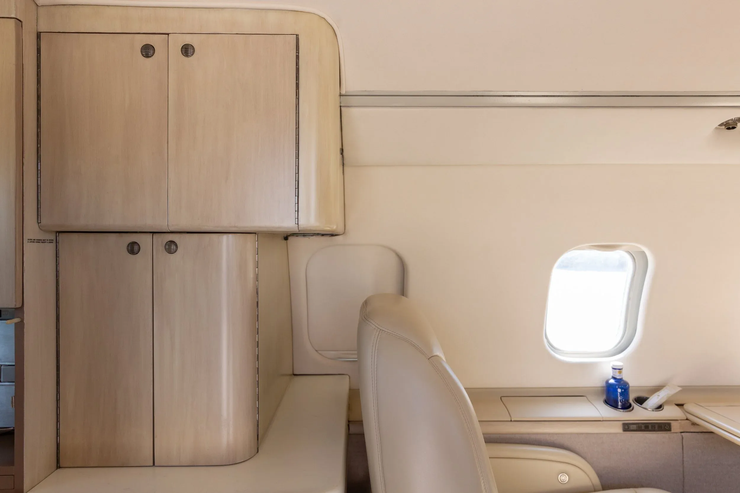 Interior view of a private jet cabin showing light wood-paneled storage cabinets and a window.