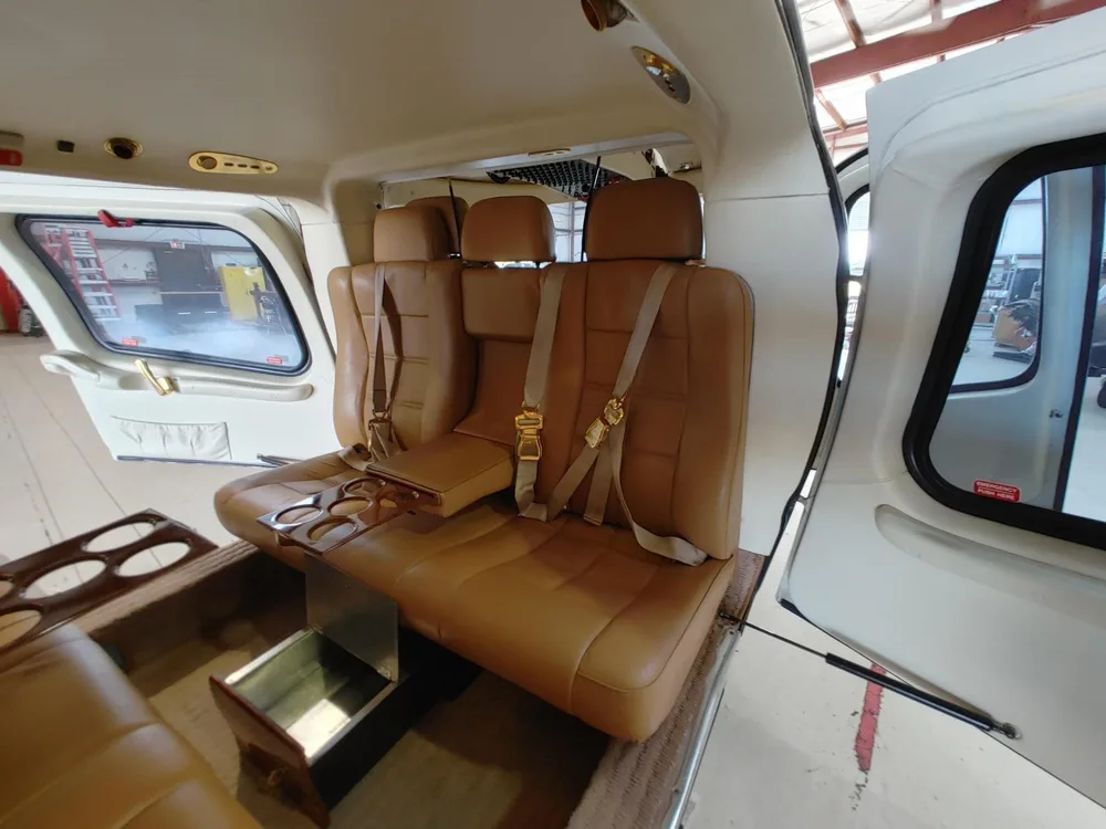 The tan leather interior cabin of a helicopter features a three-seat bench with cup holders and an open door.