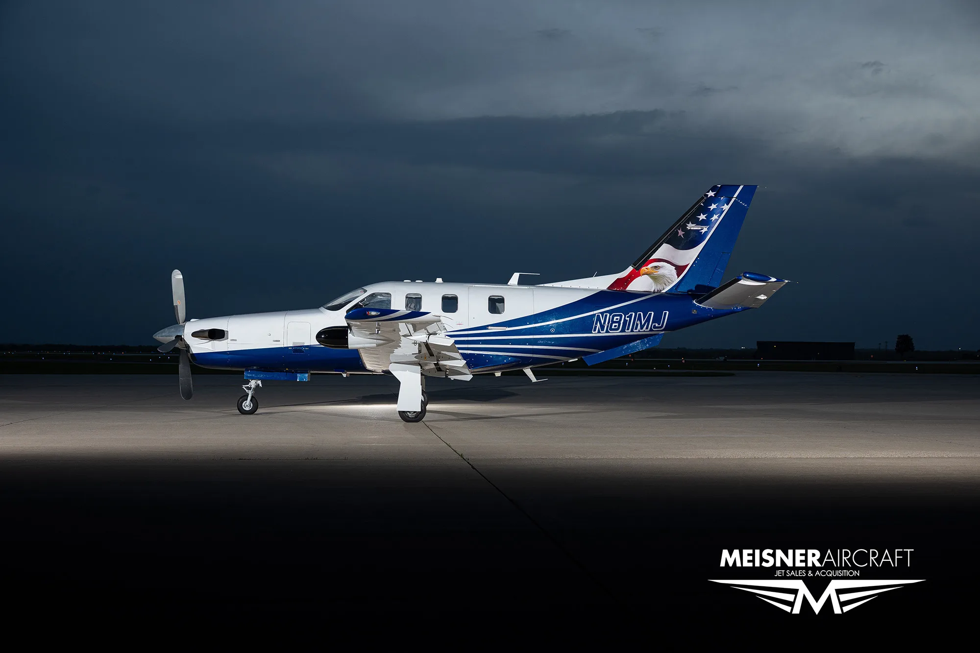 A TBM turboprop aircraft with a blue and white paint scheme featuring an eagle on the tail, parked on an airfield at dusk.