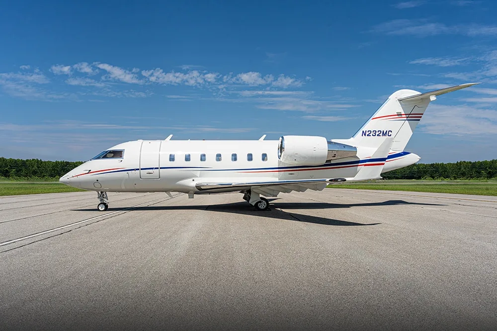 A white Bombardier Challenger business jet with red and blue stripes is parked on an asphalt runway under a clear blue sky.