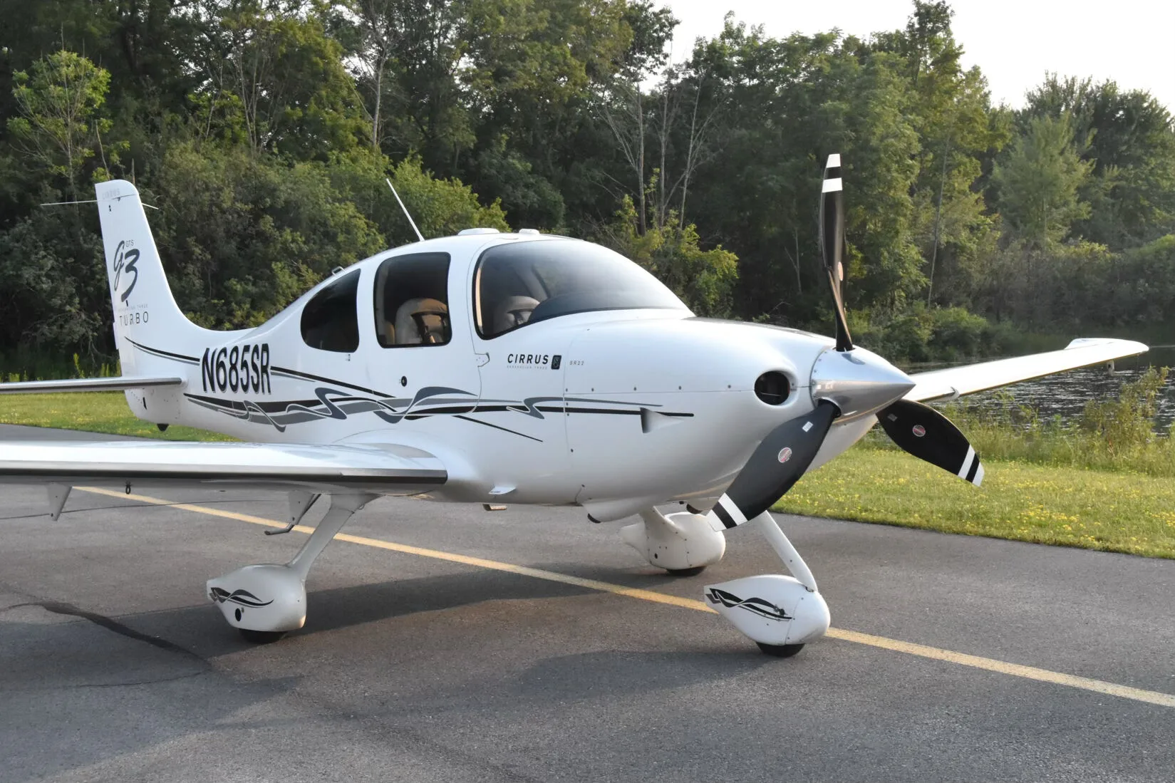 A front-quarter view of the white Cirrus SR22T aircraft (N685SR) parked on an asphalt surface with trees in the background.