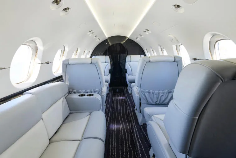A view down the aisle of the business jet's interior cabin, showing the back of the light grey leather seats.