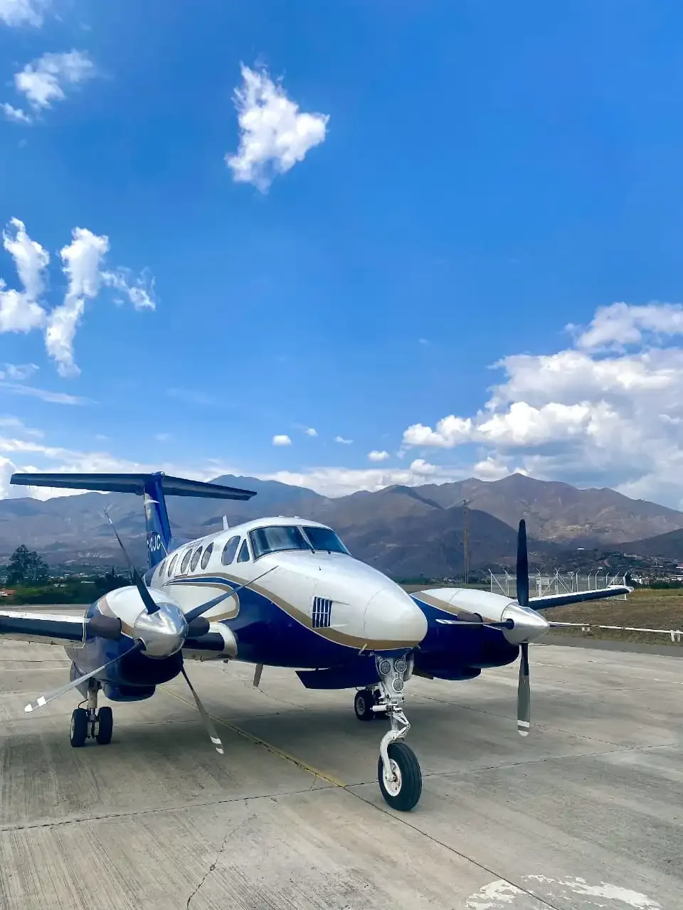 A blue and white Beechcraft King Air HC-CJC parked on an apron with mountains under a blue sky.