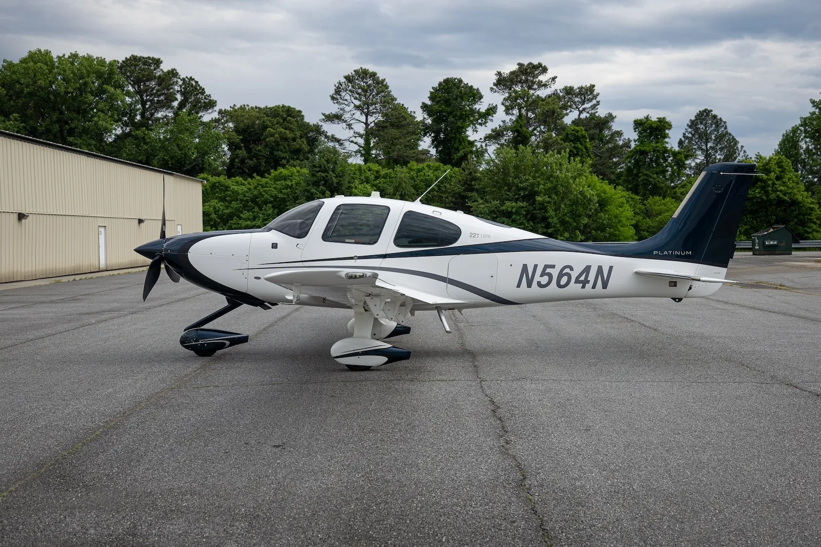 A white and dark blue Cirrus SR22T aircraft with registration N564N parked on an asphalt ramp with trees in the background.