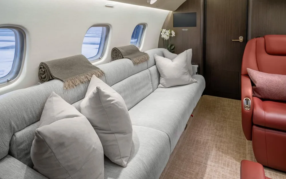 A long grey sofa with pillows and blankets next to windows in a private jet cabin, with a red leather seat visible on the right.
