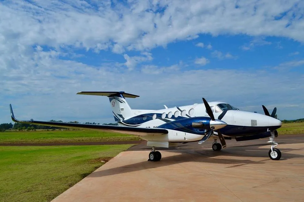 A white and blue Beechcraft King Air twin-turboprop aircraft parked on a tarmac under a partly cloudy sky.