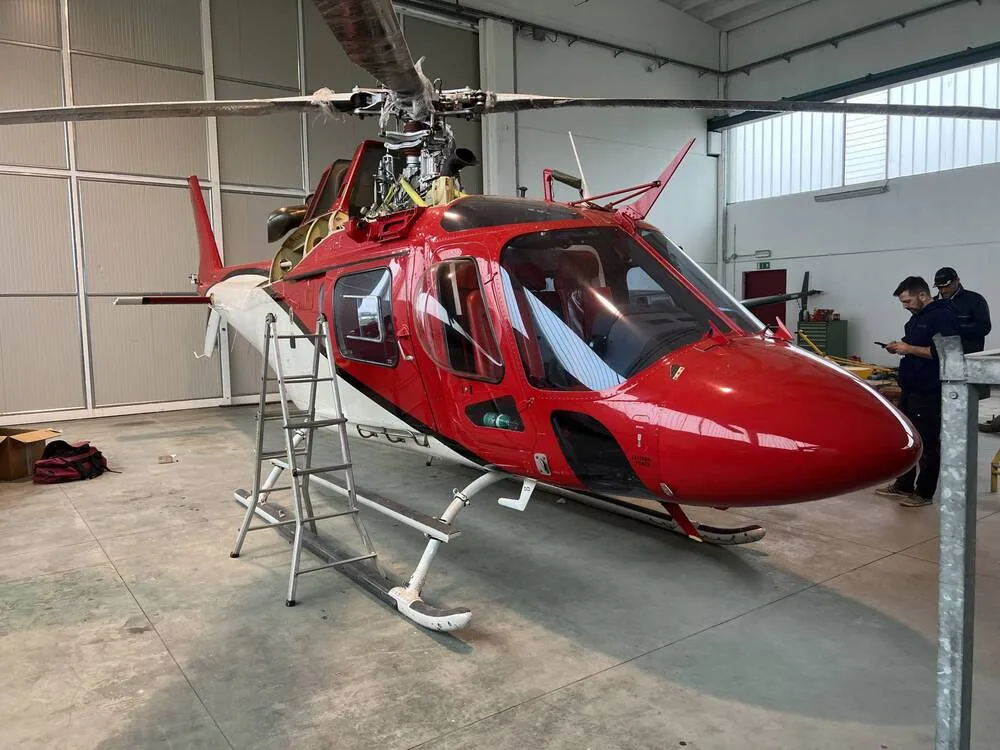 A red and white helicopter with its main rotor blades extended, parked in a hangar with people nearby.