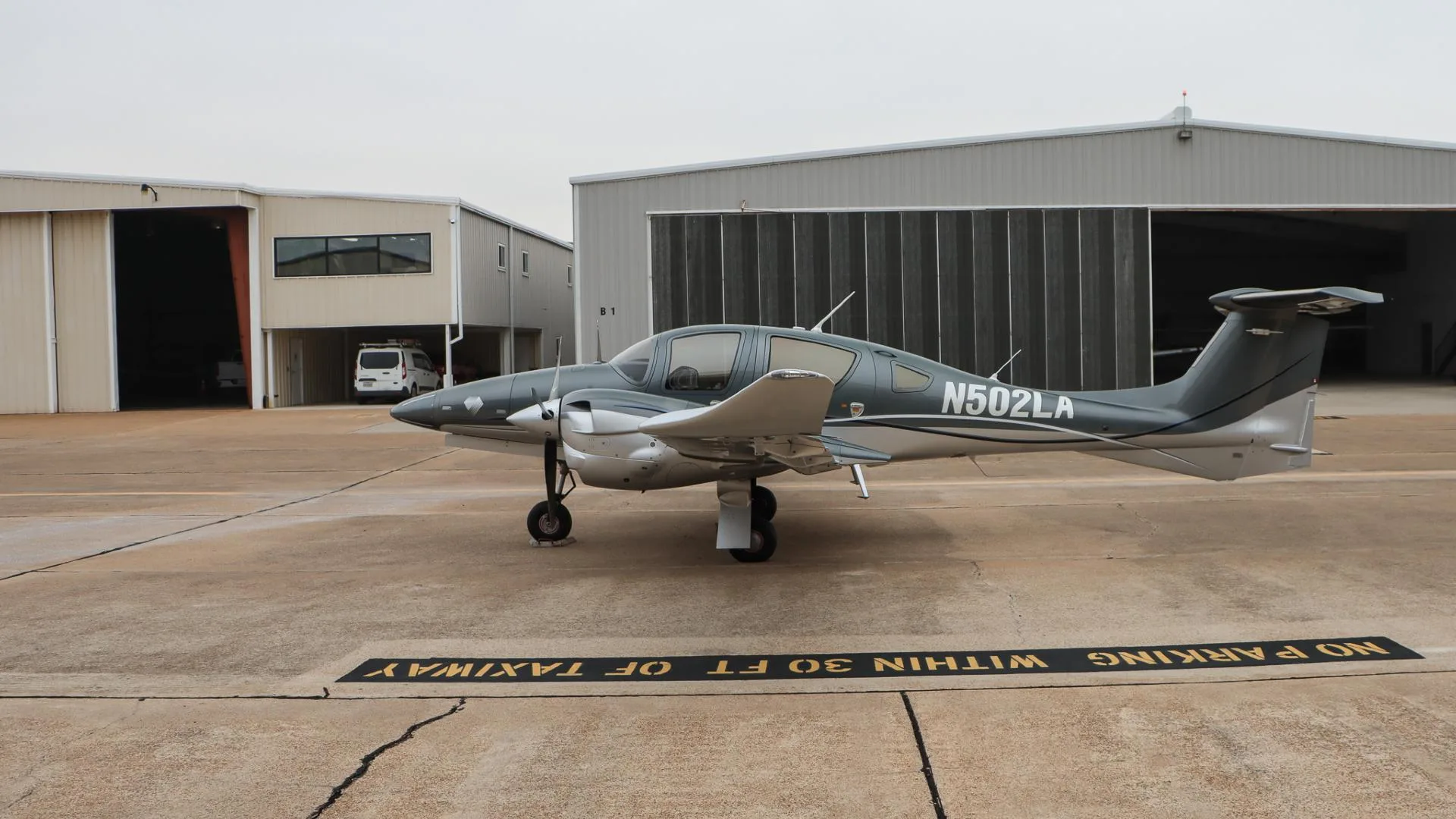 A gray and silver Diamond DA62 aircraft, N502LA, parked on an airport tarmac with hangars in the background.