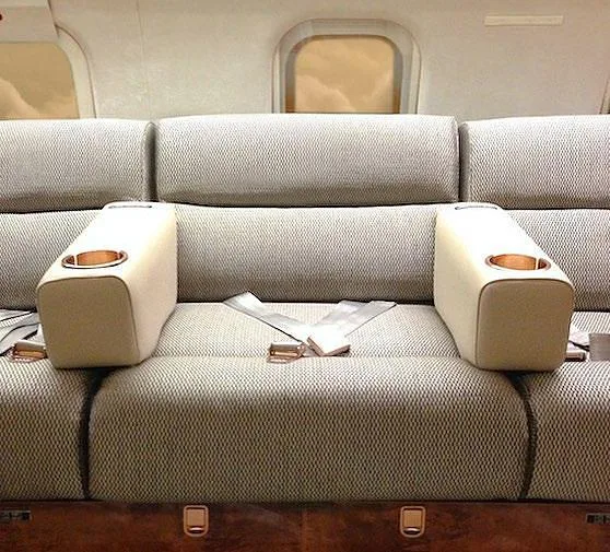 A close-up of a beige fabric bench seat in a private jet cabin, featuring two armrests with cup holders.