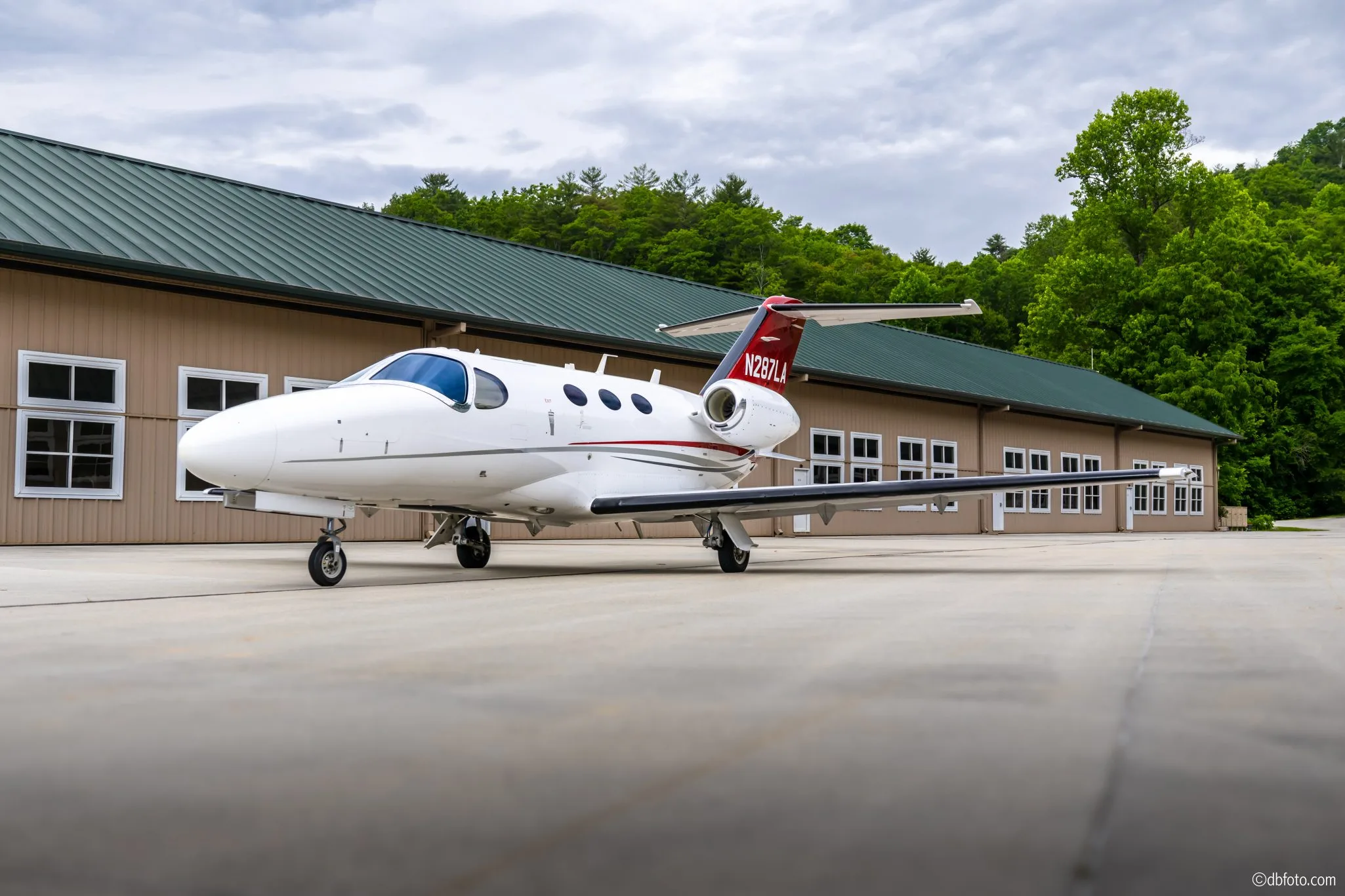 A white Cessna Citation Mustang business jet with red and grey accents parked on an tarmac in front of a hangar.