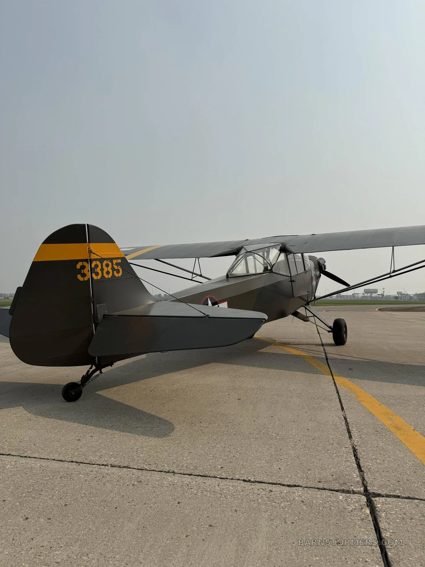 A military green Piper Cub-like aircraft with yellow tail markings and a star insignia, parked on an asphalt runway.