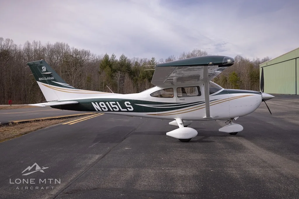 A side view of a white and dark green Cessna Skylane N915LS with gold stripes and flaps extended, parked on an asphalt ramp.