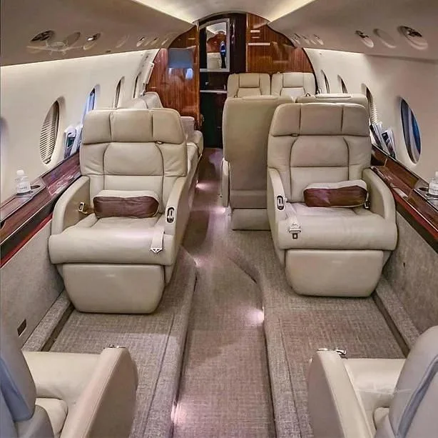 A view down the aisle of a business jet's cabin, showing rows of comfortable beige leather seats and a plush carpet.