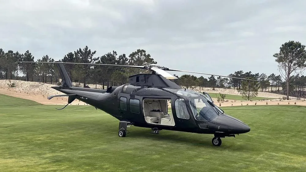 A dark grey helicopter with its passenger door open, revealing a light-colored interior, parked on a green golf course.
