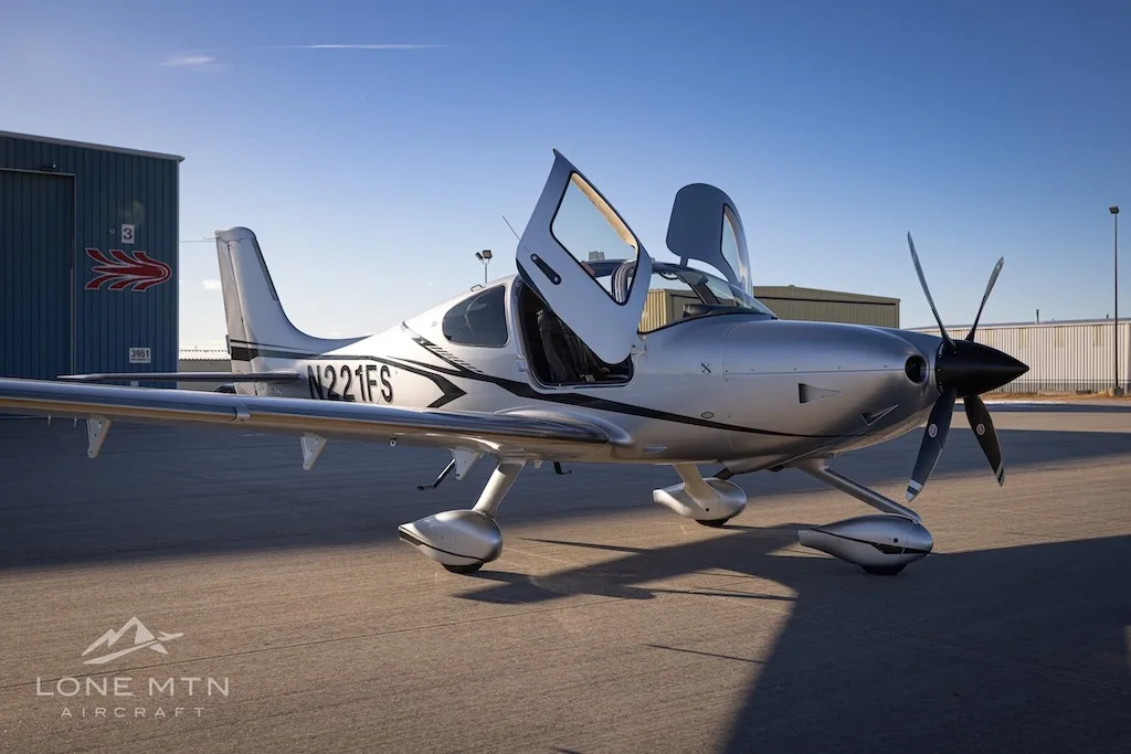 A three-quarter front view of the silver Cirrus SR22T aircraft N221FS with its doors open, parked on an airfield.