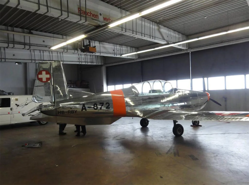 A rear-quarter view of a polished silver and orange Pilatus P-3 aircraft with Swiss markings, parked inside a hangar.