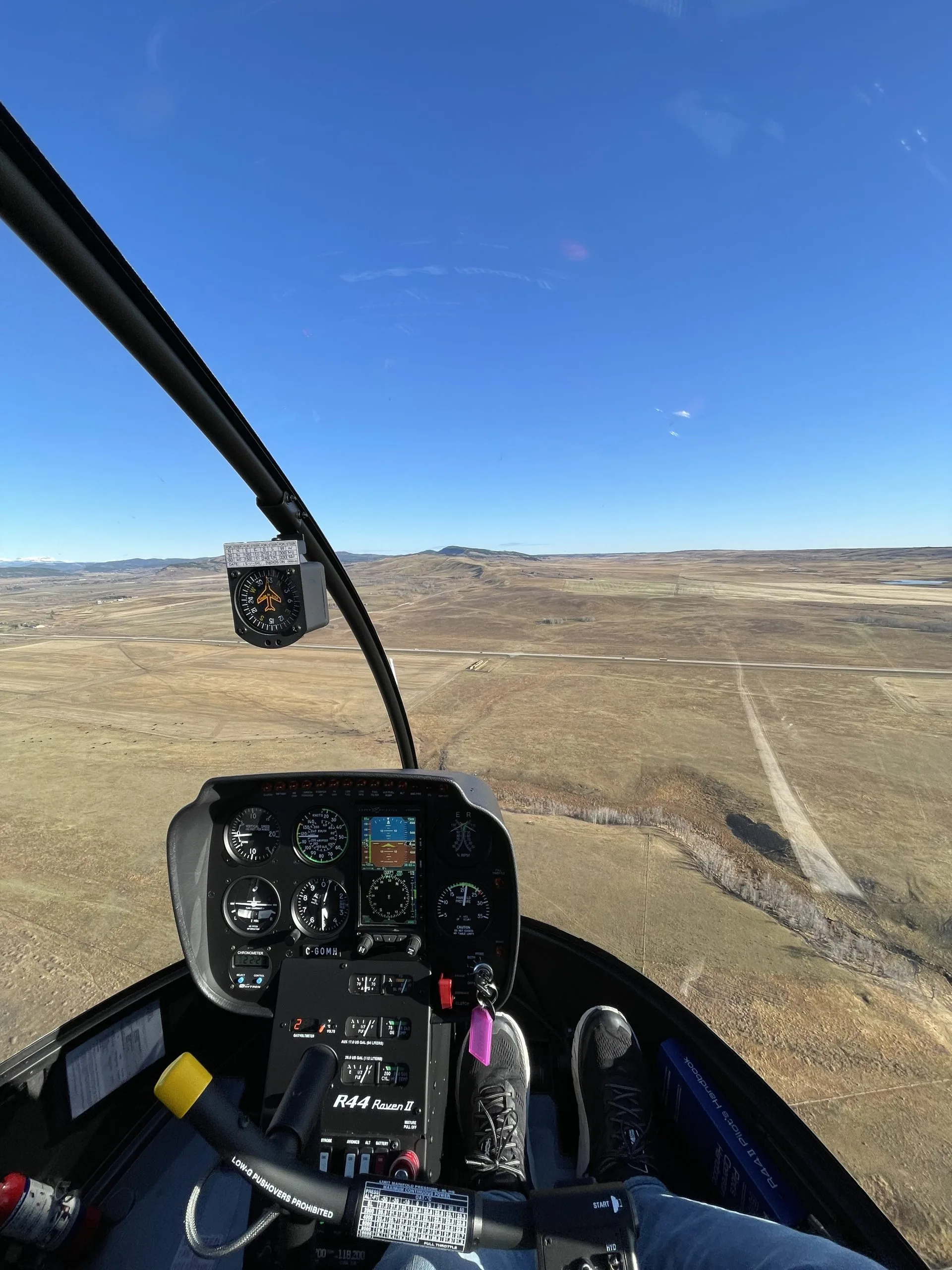 A pilot's view from the cockpit of a Robinson R44 Raven II helicopter, showing the instrument panel and a vast, dry landscape with a road below.