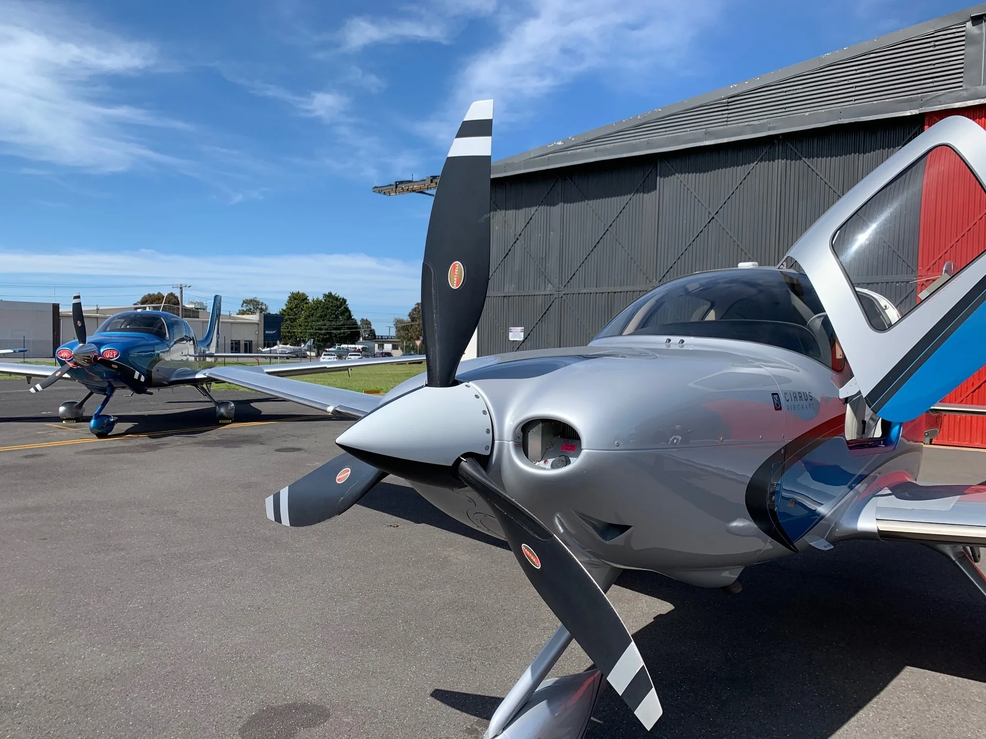 A close-up front view of a silver and blue Cirrus SR22T aircraft with open doors, parked on an airfield next to a hangar.