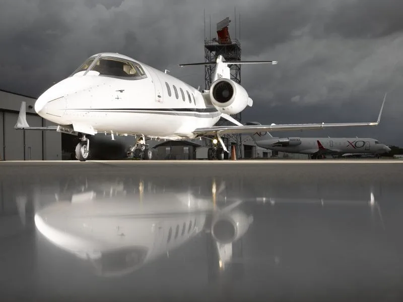 A white Learjet 60, registration N60GF, parked on a wet tarmac with a radar tower and another aircraft in the background under a cloudy sky.
