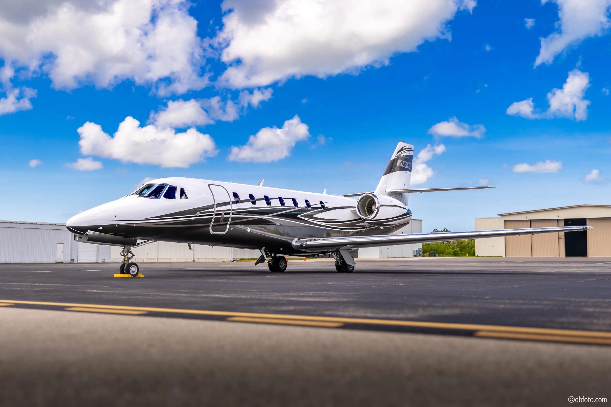 A white Cessna Citation business jet with black and silver swirl paint scheme, parked on an airfield under a bright blue sky with white clouds.