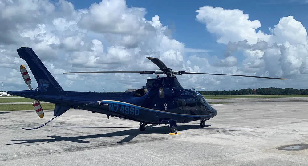 A dark blue Leonardo AW109 helicopter with gold stripes is parked on an airfield, viewed from the rear-left.