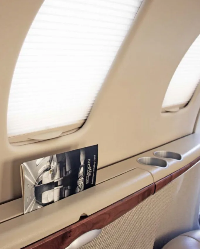 A view of an aircraft cabin interior showing a window with a shade, a side panel with cup holders, and a brochure.