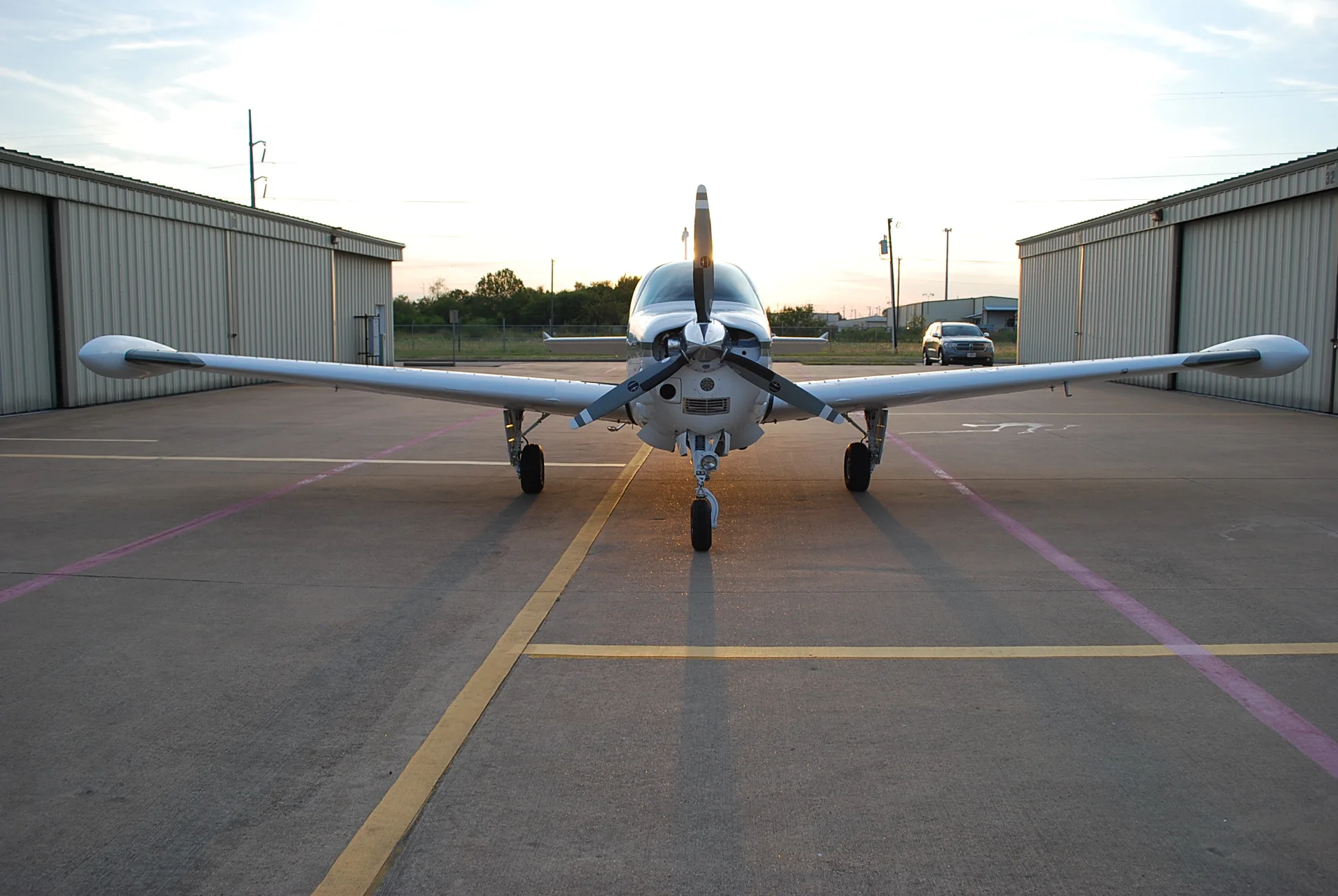 A white single-engine propeller aircraft is parked head-on on an airport tarmac with hangars in the background at sunset.