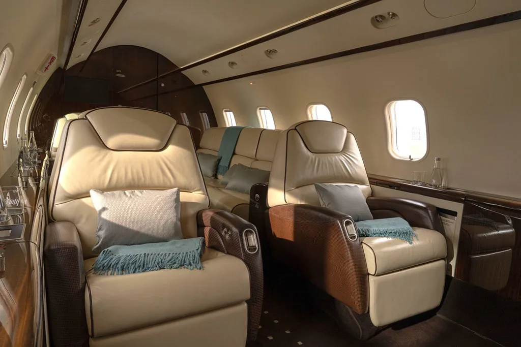 A side view of the private jet's cabin, showing two beige leather seats with blue throws and pillows, and a dark wood side panel with drinks.