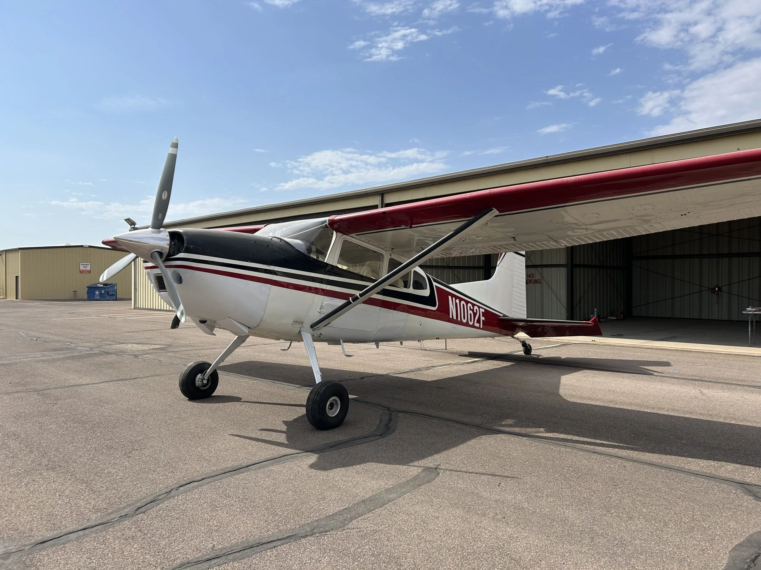 A white, black, and red Cessna 180 aircraft (N1062F) parked on an airport tarmac next to a hangar.
