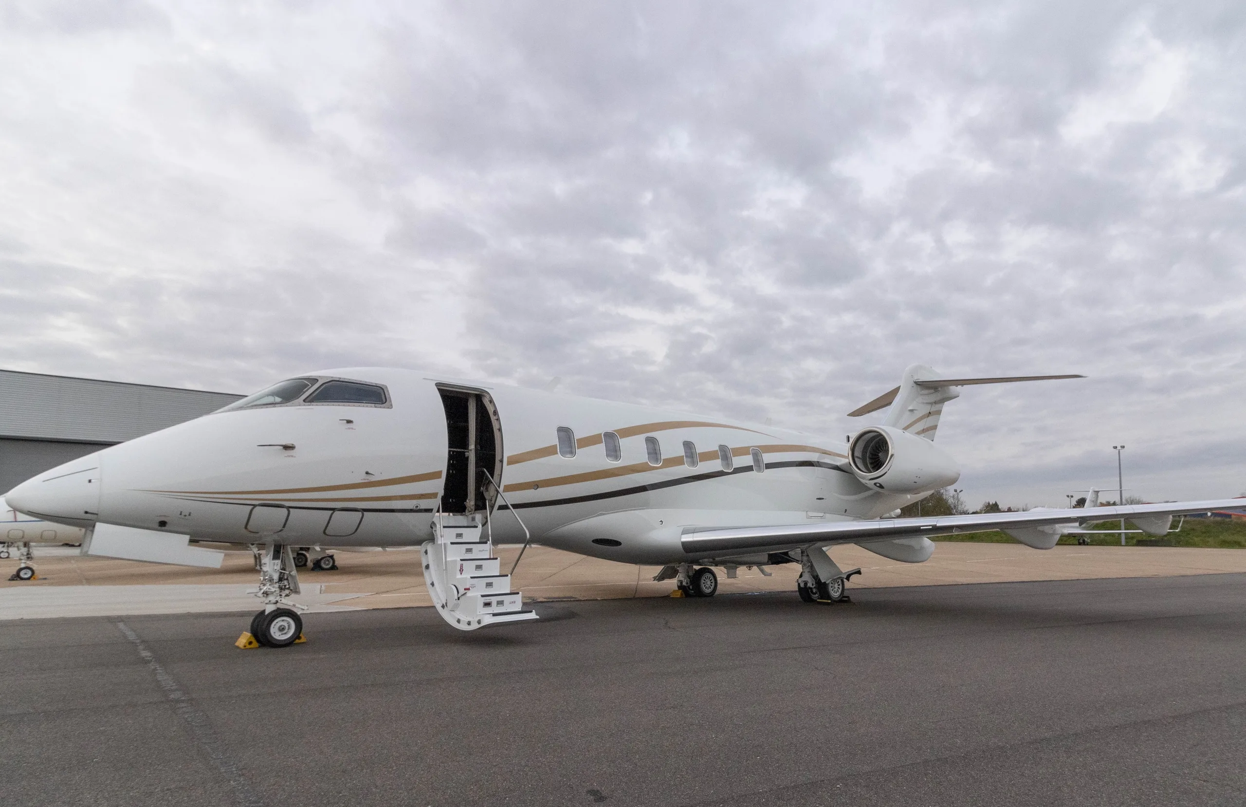 A white Bombardier Challenger 350 business jet with gold and black stripes is parked on an apron with its main door open and stairs extended under a cloudy sky.