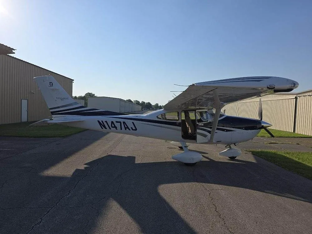 Side view of a white and blue Cessna 182 aircraft with registration N147AJ and an open door, parked on an asphalt ramp.